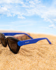 A photo of black sunglasses with blue temples.open on some sand with a blue sky in background
