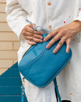 A woman holding a cubed-shaped bag in a vivid blue color by her hip, holding the strap about to open it