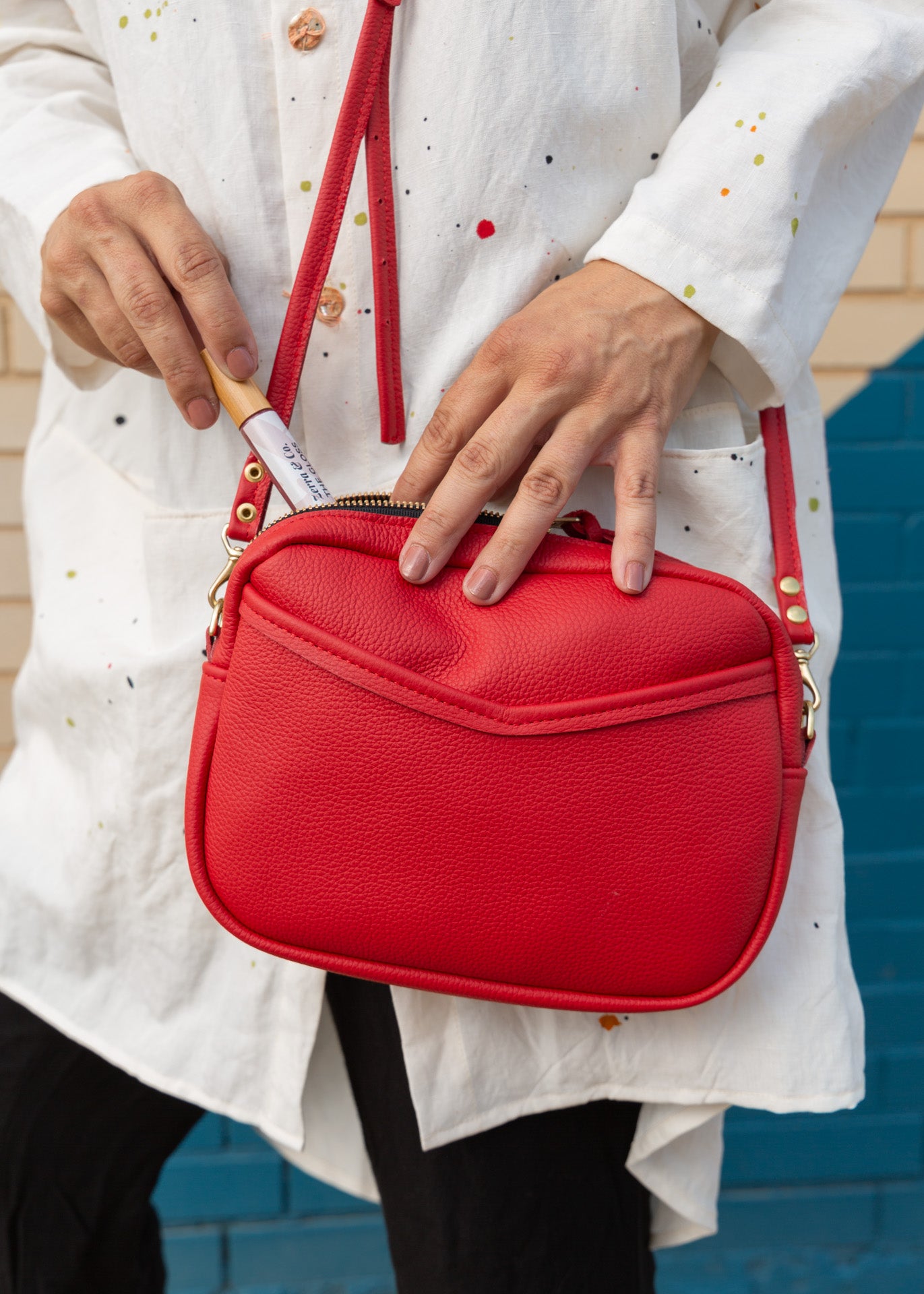A woman holding a vivid red, cherry-colored cube-shaped bag, putting a zerra &amp; co lipgloss into the crossbody, shoulder bag