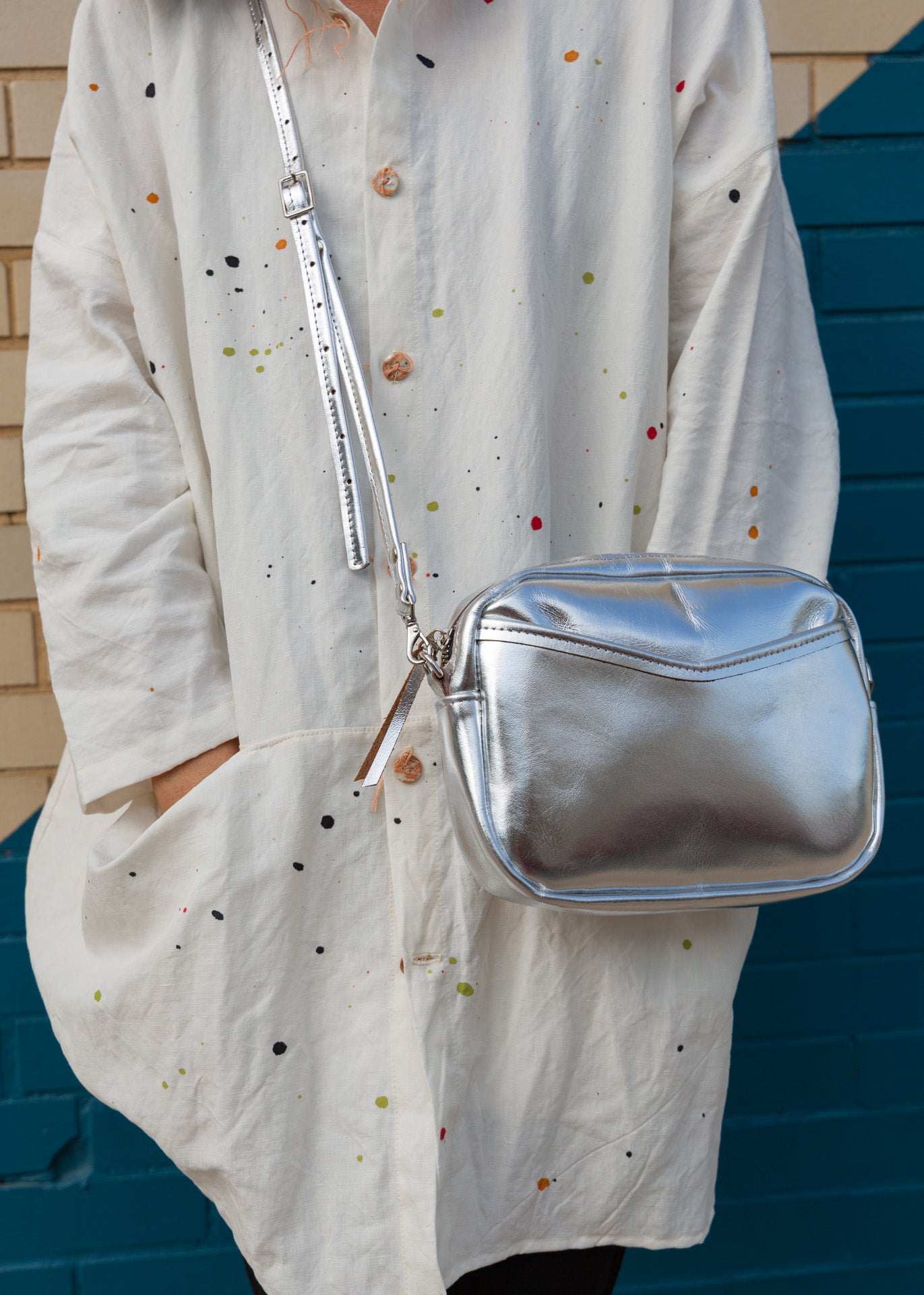 A woman in front of a brick wall with a silver cubed shaped crossbody bag sitting by her hip.