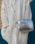 A woman in front of a brick wall with a silver cubed shaped crossbody bag sitting by her hip.