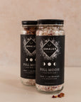 Two tall jars with black labels with one skewed behind the other and a few dried roses and salt in front of them