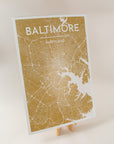 An art print of a map of Baltimore in the color golden yellow, sitting on an art easel with a light pink background