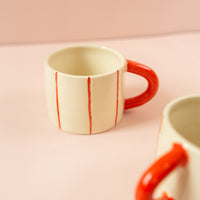 Photo of a cream ceramic mug with light red stripes going around the mug vertically, and a red handle. Another mug is in the foreground