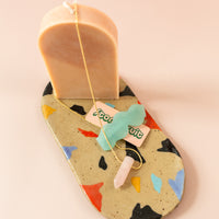 Oval-shaped ceramic tray with funky shapes painted around in various colors from blue to red to pink to black. On the tray there is a pink candle, blue hairclip and rose quartz necklace
