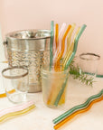 a jar of colorful wavy reusable straws with straws laid throughout, glasses surrounding and an ice bucket in the background