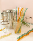 a jar of colorful wavy reusable straws with straws laid throughout, glasses surrounding and an ice bucket in the background