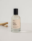 Perfume bottle on a white background with two sticks on palo santo in the background with a light pink label saying "Palo Santo"
