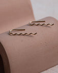 Two gold wavy earrings on a wavy display object with a neutral background