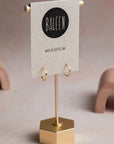 Gold earrings on a stand with 'Baleen' brand card on a beige background