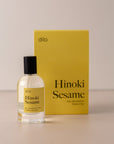 Bottle of perfume by dilo, with a yellow label on the front stating "hinoki sesame. eau de parfum, 50 ml. made in the usa" with the yellow box that it comes in in the background