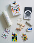 Collection of items including a prayer candle, oracle cards, palo santo, sage and matches on a light surface