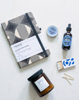 Collection of items including a notebook, candle, tincture, salve and matches on a light surface.