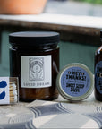 Collection of wellness products including a notebook, candle, tincture, salve, and matches on a table outdoors.
