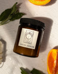 flat lay of a candle labeled "lucid dream" with orange halves and sage besides the candle on a white background