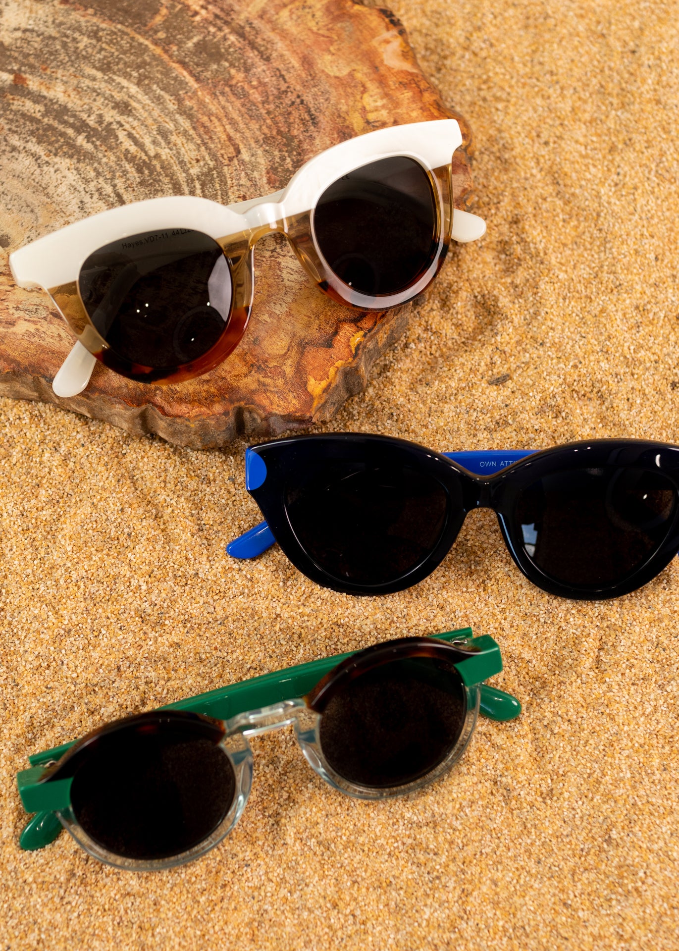 A photo of three types sunglasses in multiple colors folded on some sand &amp; a tan rock piece