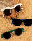 A photo of three types sunglasses in multiple colors folded on some sand & a tan rock piece