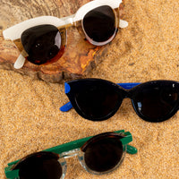A photo of three types sunglasses in multiple colors folded on some sand & a tan rock piece