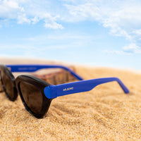A photo of black sunglasses with blue temples.open on some sand with a blue sky in background