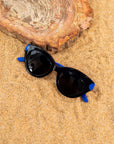 A photo of black sunglasses with blue temples. folded on some sand