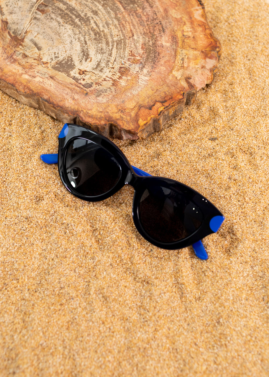 A photo of black sunglasses with blue temples. folded on some sand