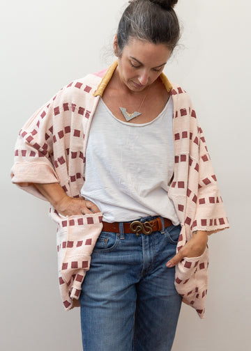 Photo of model wearing a pink jacket that goes to mid thigh, in the color pink with lines of dark pink square patters all over. Jacket has a mustard yellow color collar detail, and is 3/4 sleeves