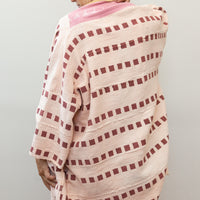 Photo of model wearing a pink jacket that goes to mid thigh, in the color pink with lines of dark pink square patters all over. Jacket has a mustard yellow color collar detail, and is 3/4 sleeves