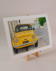 Art print of a vintage, yellow fiat on the side of the road in italy, standing on an easel with a light pink background