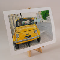 Art print of a vintage, yellow fiat on the side of the road in italy, standing on an easel with a light pink background