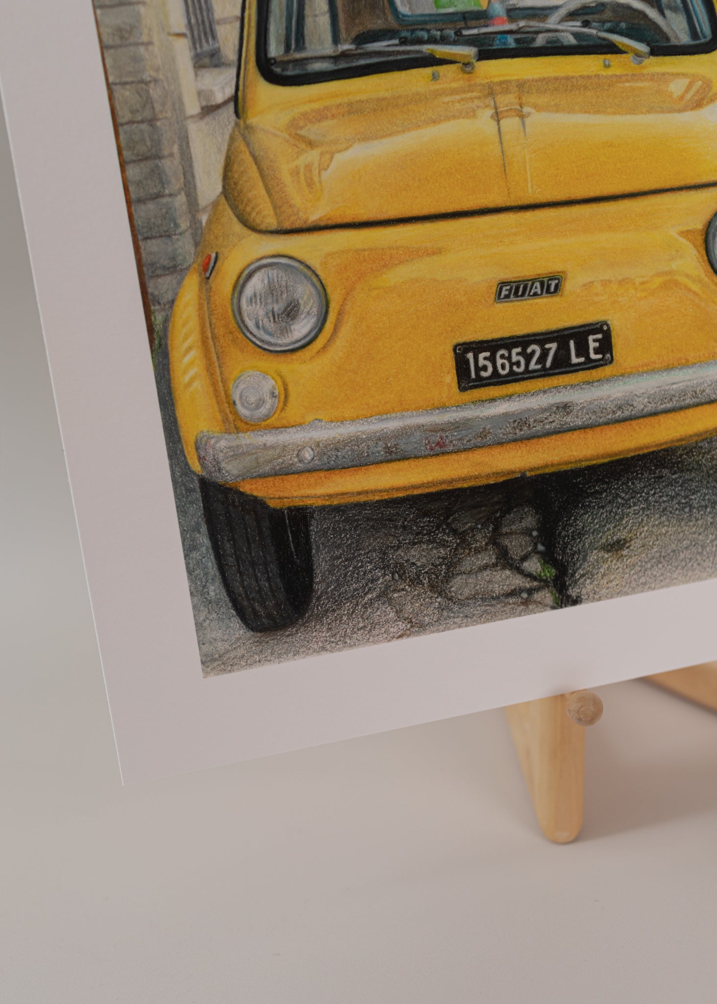 Close-up of art print of a vintage, yellow fiat on the side of the road in italy, standing on an easel with a light pink background