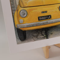 Close-up of art print of a vintage, yellow fiat on the side of the road in italy, standing on an easel with a light pink background