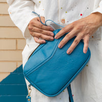 A woman holding a cubed-shaped bag in a vivid blue color by her hip, holding the strap about to open it