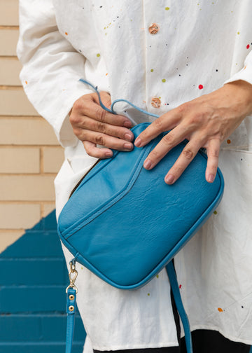 A woman holding a cubed-shaped bag in a vivid blue color by her hip, holding the strap about to open it