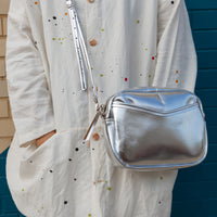 A woman in front of a brick wall with a silver cubed shaped crossbody bag sitting by her hip.