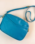 Flatlay of cubist handbag in azure, or blue, with a v-shaped front pocket and a long thin shoulder strap