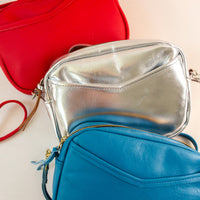 Layer of cubist handbags, with a v-shaped front pocket and a long thin shoulder strap, in cherry, silver and azure