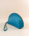 Azure, or blue, colored shell bag standing up on a light pink background with a short wrist handle