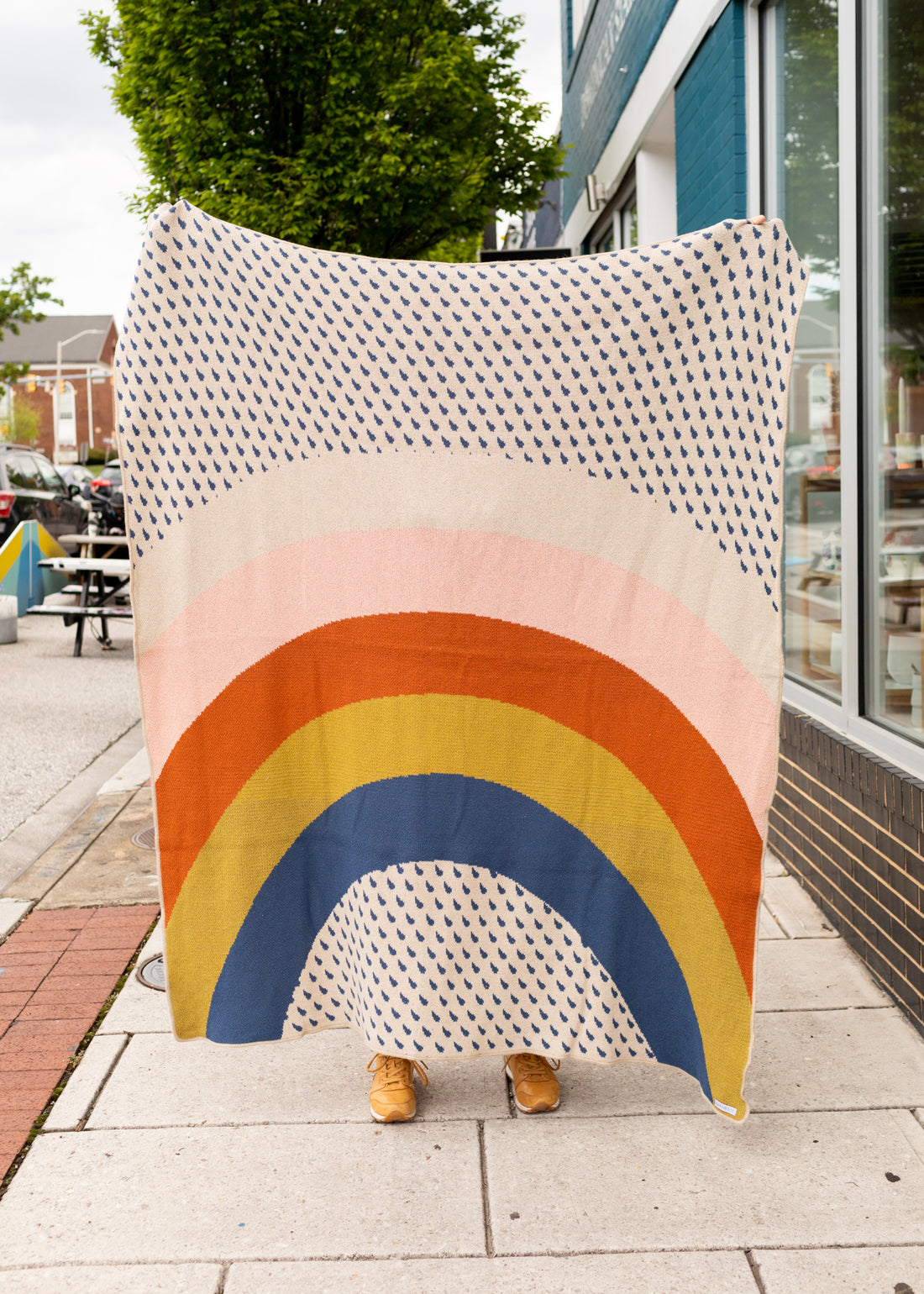 A woman holding up a blanket outside with blue raindrop dots throughout and a big rainbow in different colors on the blanket