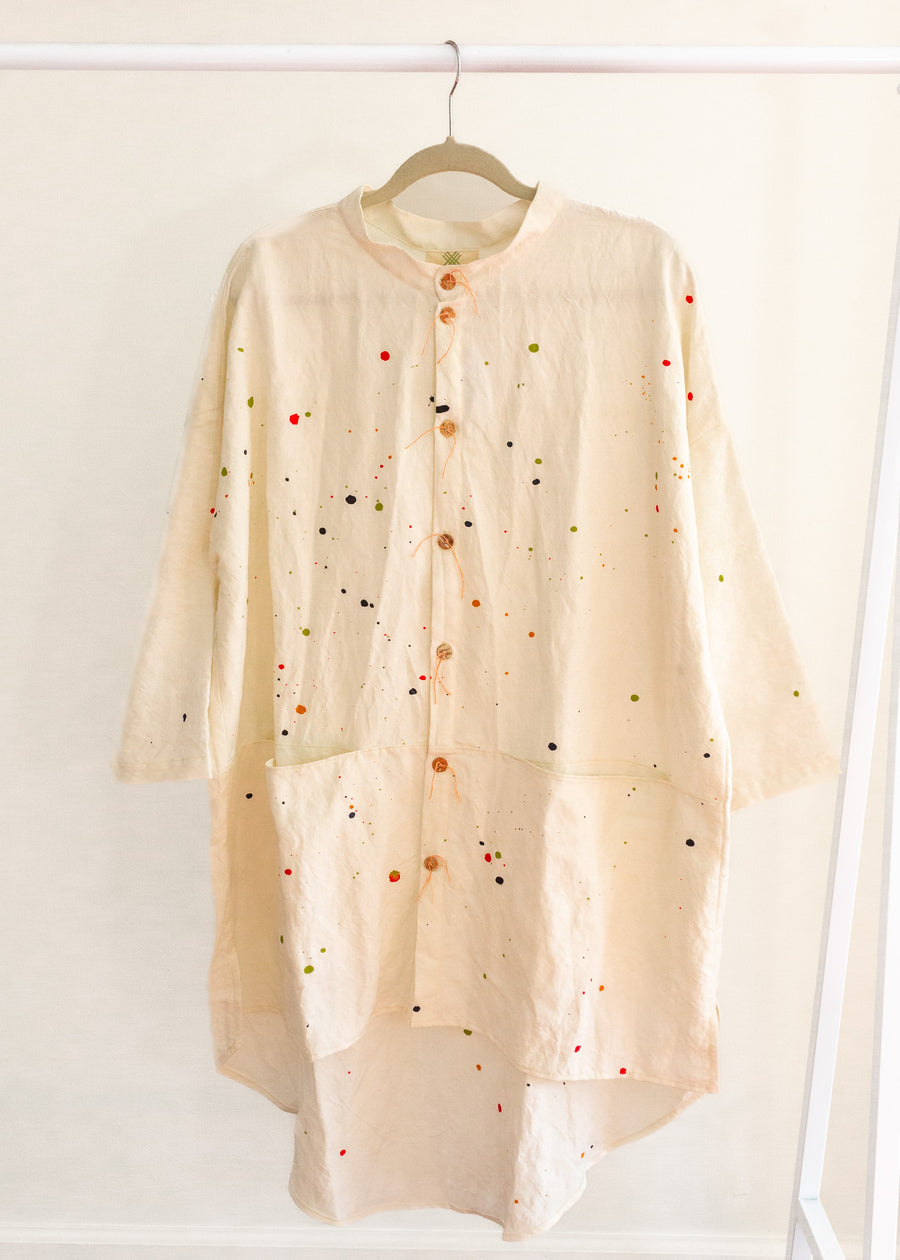 Cream colored long button-down shirt with 3/4 sleeves and paint splatter, hanging on a clothing rack.