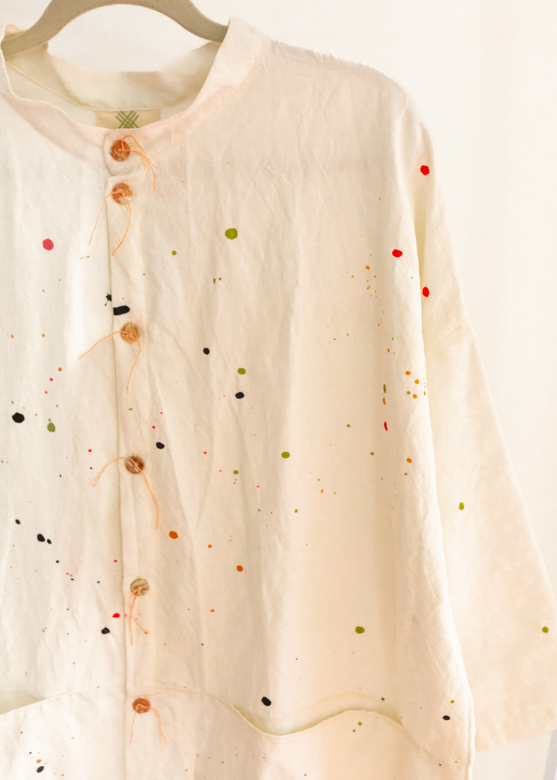 Close-up of cream colored long button-down shirt with 3/4 sleeves and paint splatter, hanging on a clothing rack.