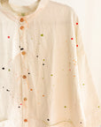 Close-up of cream colored long button-down shirt with 3/4 sleeves and paint splatter, hanging on a clothing rack.
