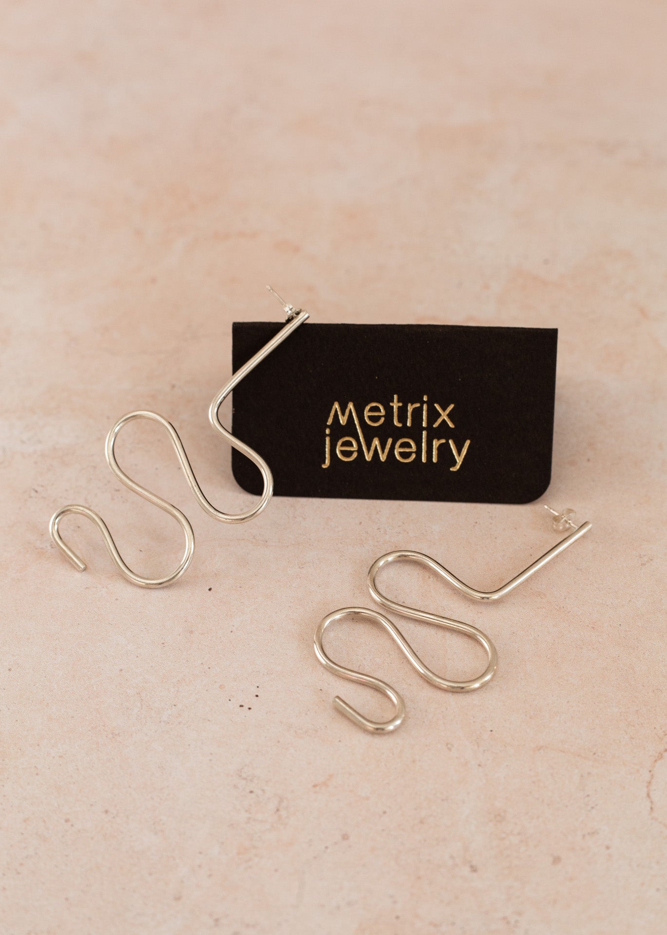 Two quiggle statement earrings in sterling silver on a light pink flatlay and a black jewelry card stating &quot;metrix jewelry&quot;