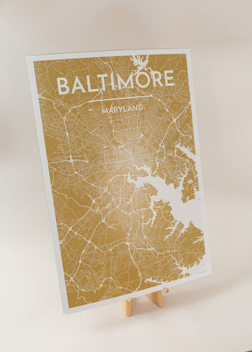 An art print of a map of Baltimore in the color golden yellow, sitting on an art easel with a light pink background