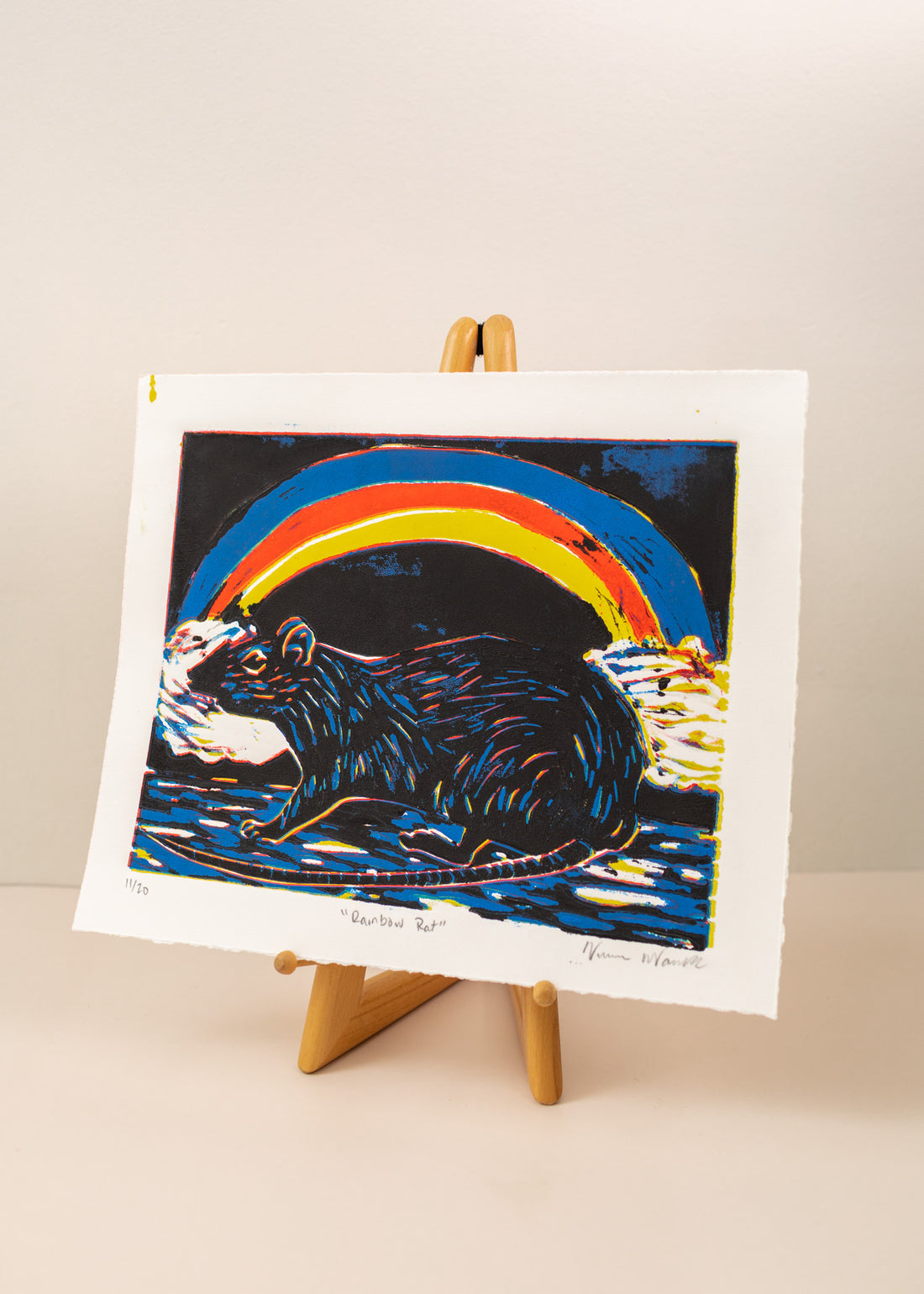 Art print of a rat with a rainbow behind it, in colors black, blue, red and yellow on a horizontal paper. Sitting on an art easel, with a light pink background
