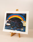 Art print of a rat with a rainbow behind it, in colors black, blue, red and yellow on a horizontal paper. Sitting on an art easel, with a light pink background