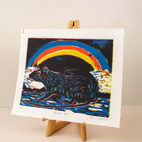 Art print of a rat with a rainbow behind it, in colors black, blue, red and yellow on a horizontal paper. Sitting on an art easel, with a light pink background