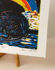 Close-up of an art print of a rat with a rainbow behind it, in colors black, blue, red and yellow on a horizontal paper. Sitting on an art easel, with a light pink background, photo is close-up of the artists signature, Virginia Warwick