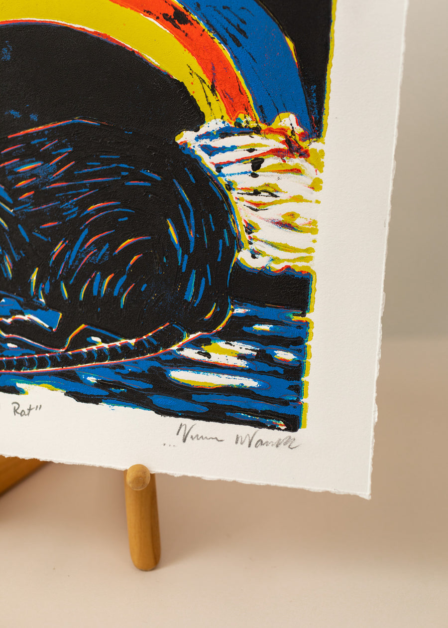 Close-up of an art print of a rat with a rainbow behind it, in colors black, blue, red and yellow on a horizontal paper. Sitting on an art easel, with a light pink background, photo is close-up of the artists signature, Virginia Warwick