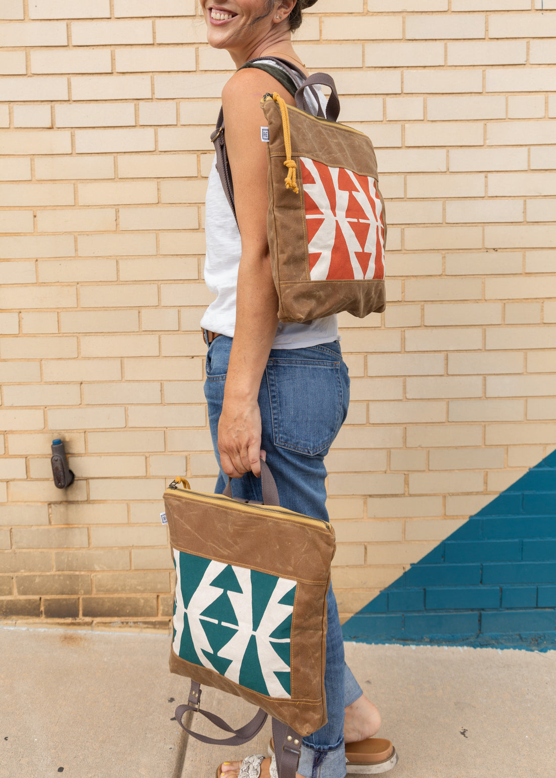 Woman holding a terra-cotta vegan leather square-shaped backpack on her left shoulder, and another one in peacock by her leg