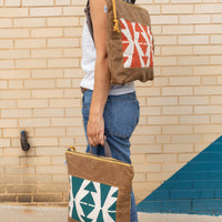 Woman holding a terra-cotta vegan leather square-shaped backpack on her left shoulder, and another one in peacock by her leg